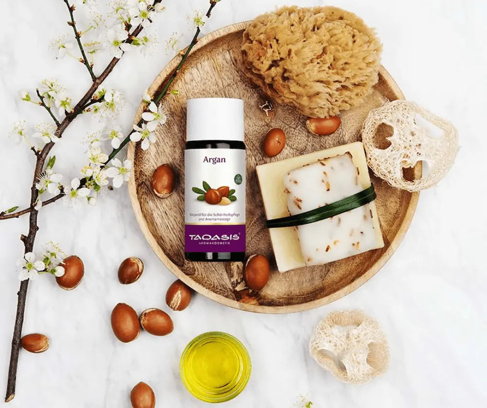 Taoasis Argan Körperöl in Flasche mit weißem Deckel und lila Etikett auf Holzteller, umgeben von Argannüssen, Seife, Schwamm und Kirschblüten