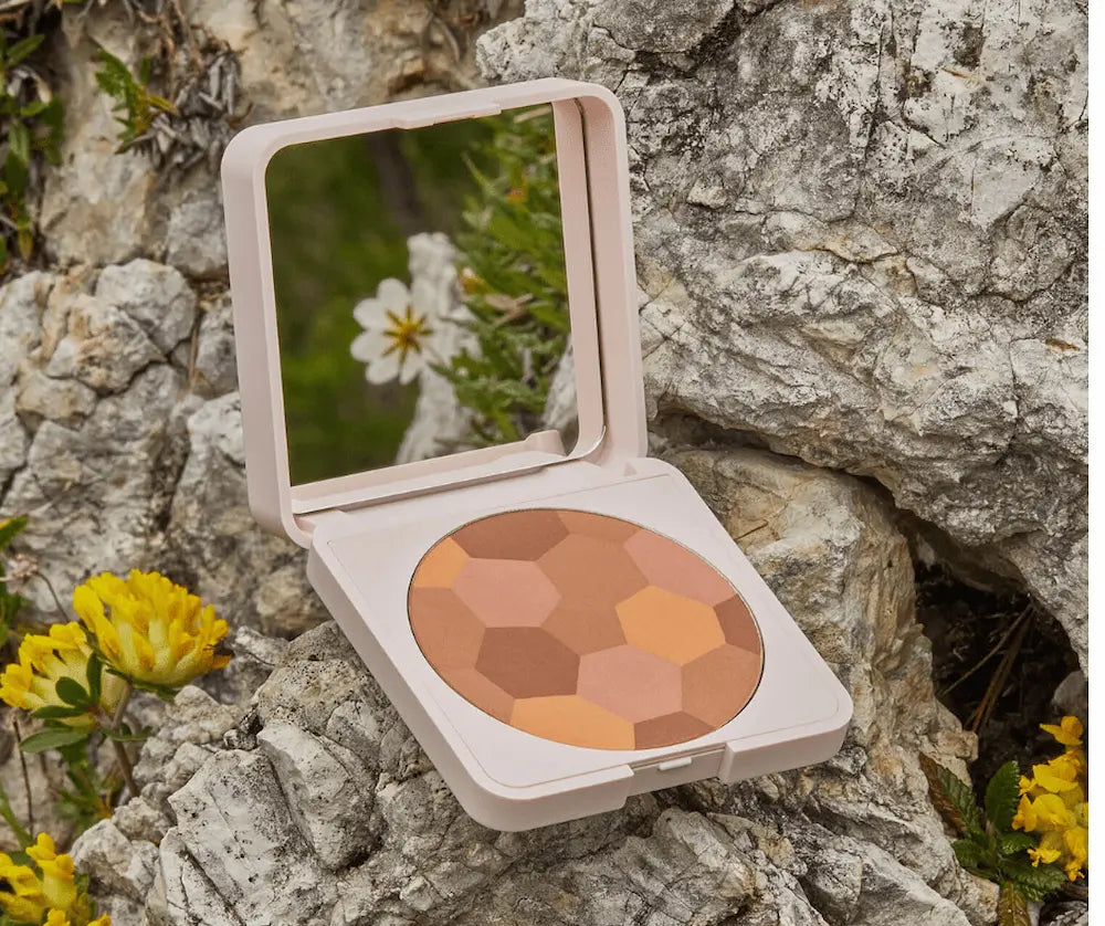 Geöffnete Puder-Kompaktdose in Rosé mit Spiegel und Mosaik-Bronzer in mehreren Brauntönen auf Felsen mit gelben Blüten.