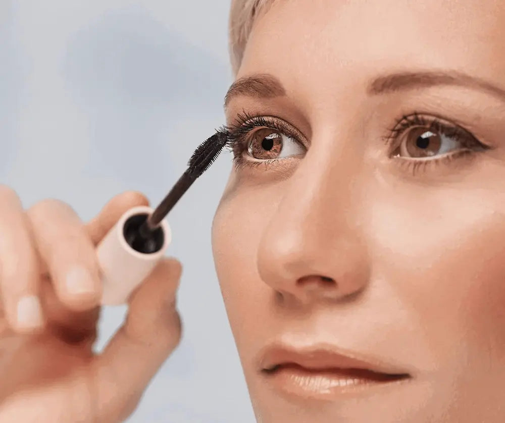 Frau trägt schwarze Mascara mit Bürstenapplikator auf die Wimpern auf, Nahaufnahme des Gesichts mit braunen Augen
