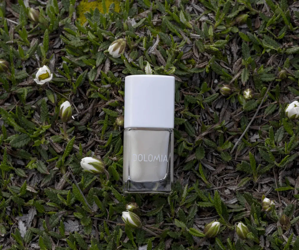 Dolomia Nagellack-Fläschchen mit weißem Deckel und milchig-weißem Inhalt, liegend auf grünem Pflanzenbett mit weißen Blütenknospen.