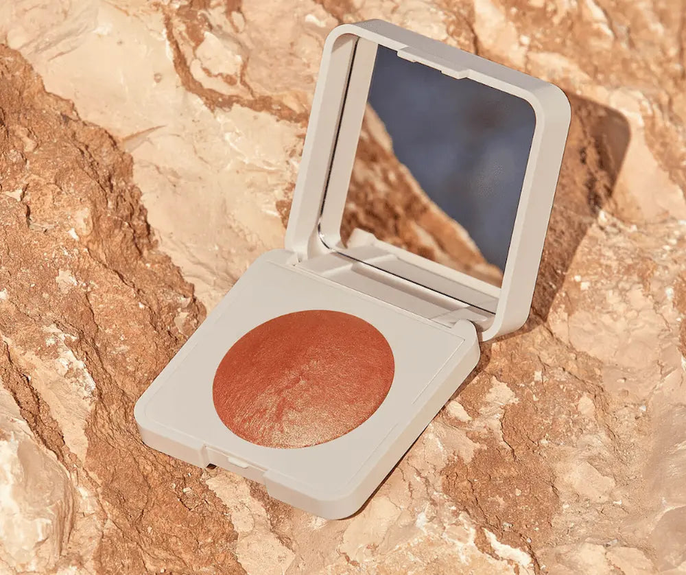 Geöffnete beige Kompaktdose mit terrakottafarbenem Rouge-Puder und Spiegel im Deckel auf hellem Natursteinuntergrund.