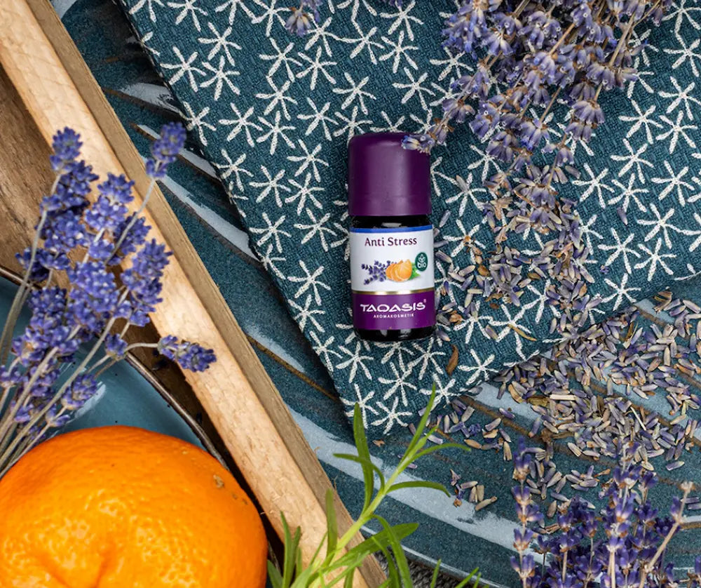 Taoasis Anti Stress Aromakosmetik Fläschchen mit violettem Deckel, umgeben von getrocknetem Lavendel und einer Orange auf blauem Tuch