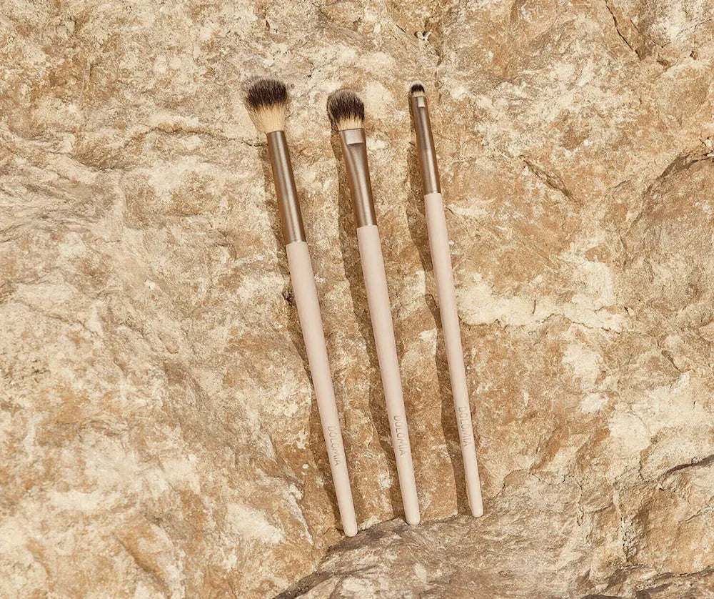Set aus drei Augen-Make-up-Pinseln mit beigen Stielen und unterschiedlichen Pinselköpfen, liegend auf hellbraunem Stein