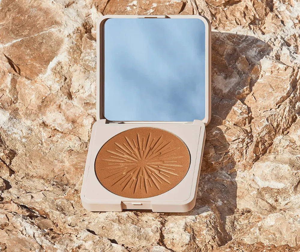 Geöffnete beige Puderdose mit Spiegel und gepresstem Bräunungspuder in Braun mit Sonnenstrahlen-Relief auf Felsuntergrund.