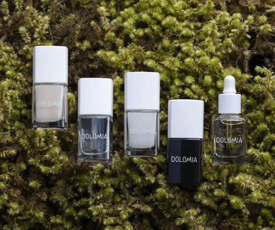 Fünf Dolomia Nagelpflegeprodukte mit weißen Verschlüssen, darunter eine Tropfflasche, auf grünem Moos angeordnet.
