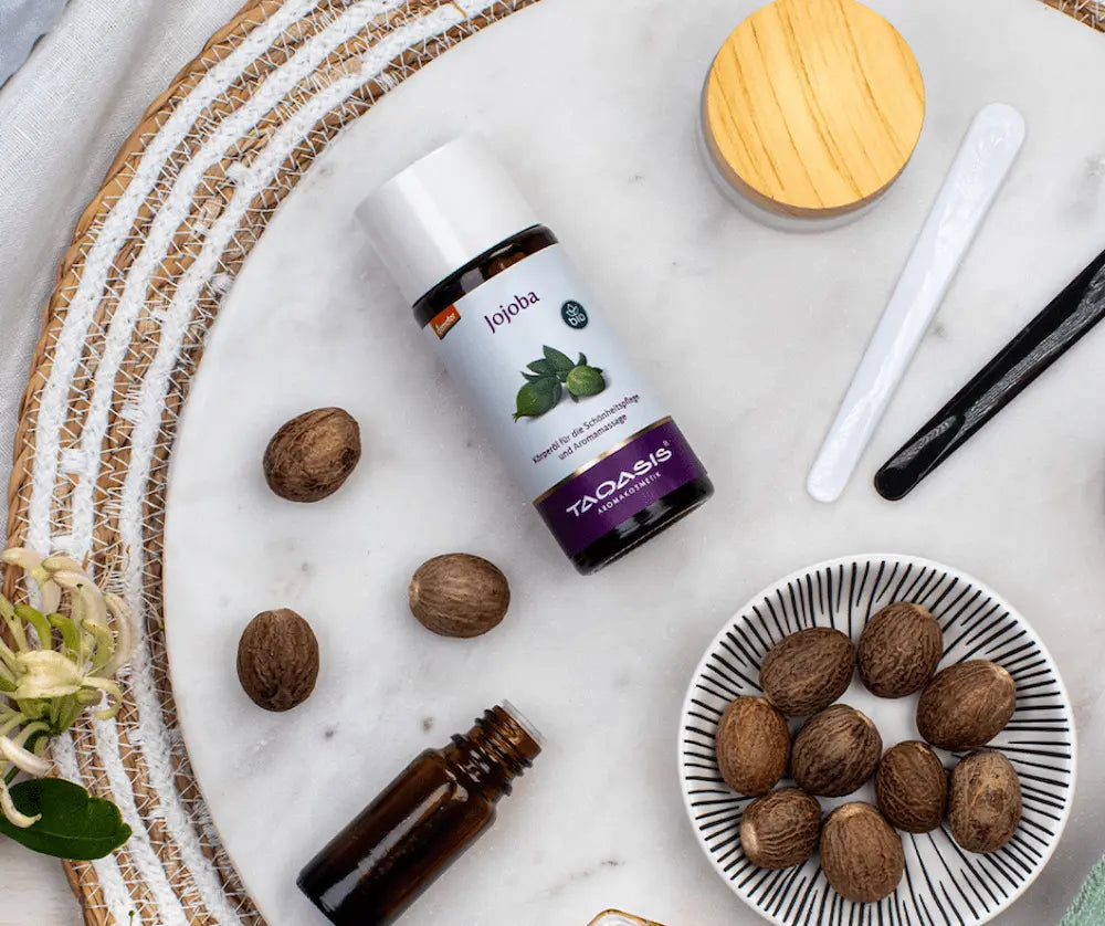 TAOASIS Jojoba Bio-Körperöl in brauner Glasflasche mit weißem Deckel auf Marmor, umgeben von Jojoba-Nüssen und Pflegespateln