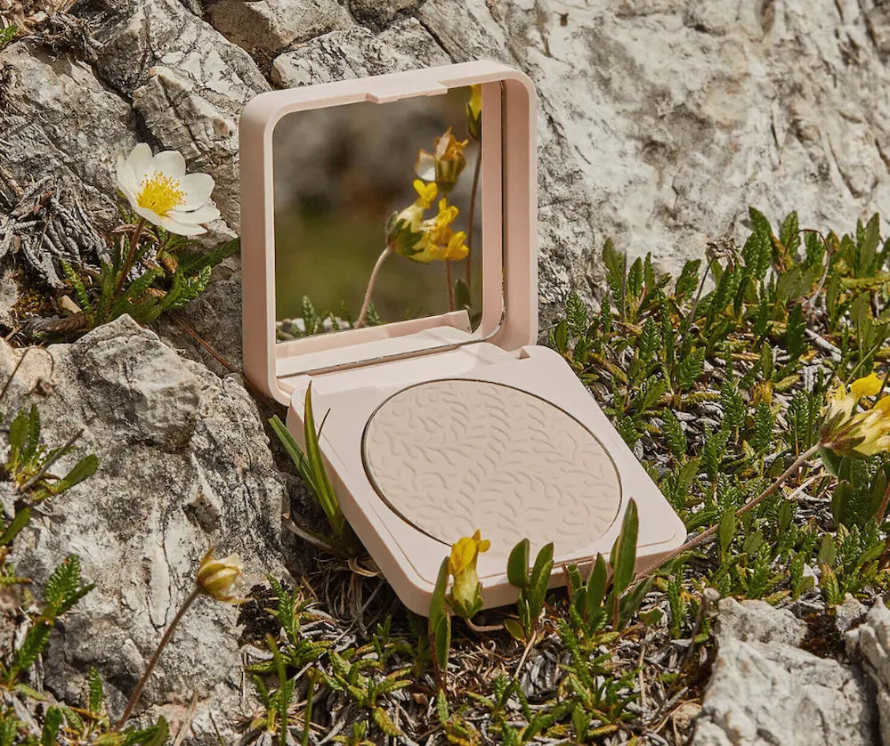 Geöffnete rosé-beige Kompaktpuder-Dose mit Spiegel und hellem Puder mit Blattprägung, platziert auf felsigem Untergrund mit Wildblumen.