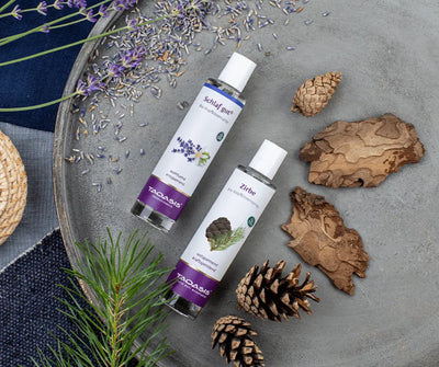 Taoasis Zirbe Bio Kopfkissenspray in weißer Sprühflasche, dahinter Schlaf gut Spray, umgeben von Zapfen, Rinde und Lavendel