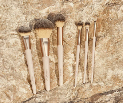 Set aus sechs Dolomia Make-up-Pinseln mit beigen Griffen und goldenen Fassungen, dekorativ auf Steinuntergrund angeordnet.