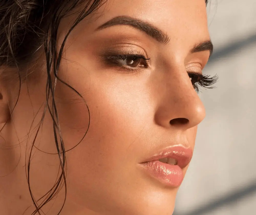 Nahaufnahme einer Frau im Profil mit dunklen, voluminösen Wimpern, natürlichem Make-up und glänzenden, rosé getönten Lippen.