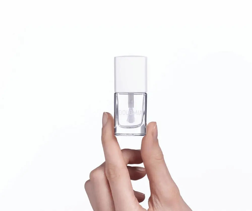 Hand hält ein kleines transparentes Nagellack-Fläschchen mit weißem Deckel und Aufschrift Dolomia vor weißem Hintergrund.