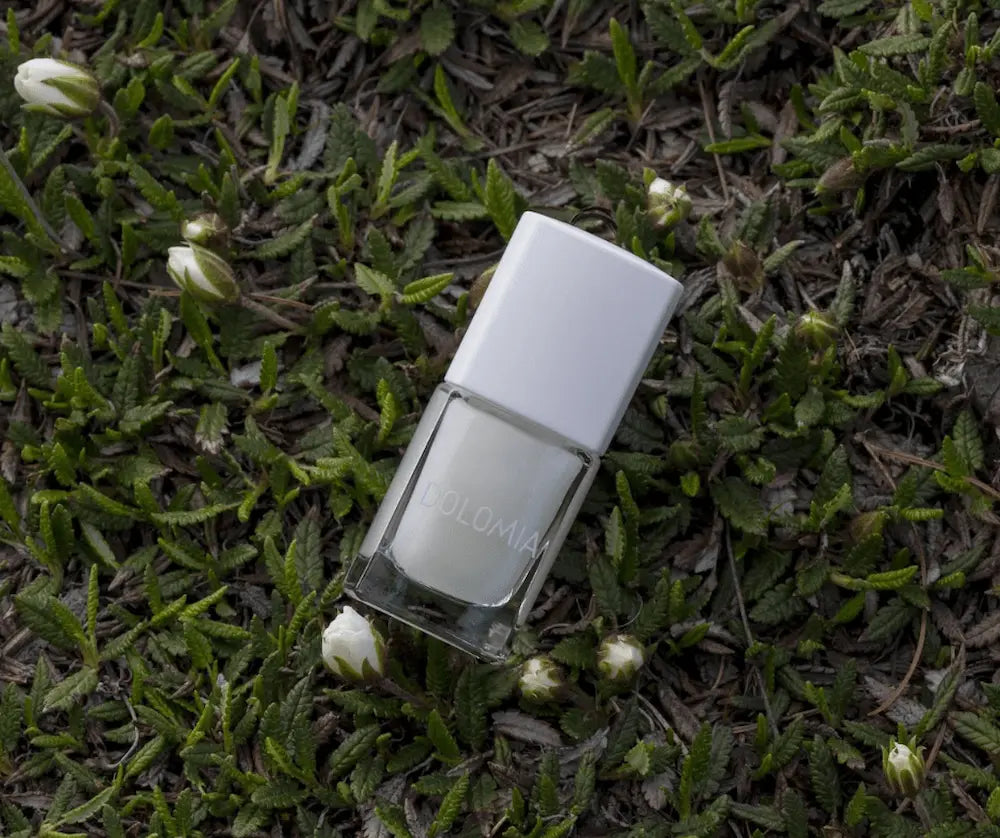 Nagellackflasche mit weißem Deckel und hellem Inhalt, liegend auf grünem Pflanzenbewuchs mit weißen Blütenknospen.