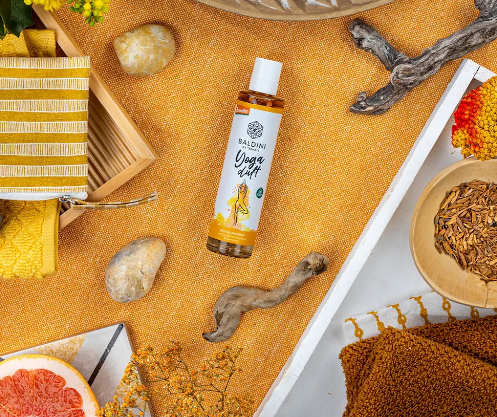 Baldini by Taoasis Yoga Duft Bio-Raumspray in schlanker Flasche mit weißem Etikett, präsentiert auf orangefarbenem Stoff mit Steinen, Wurzeln und Grapefruit