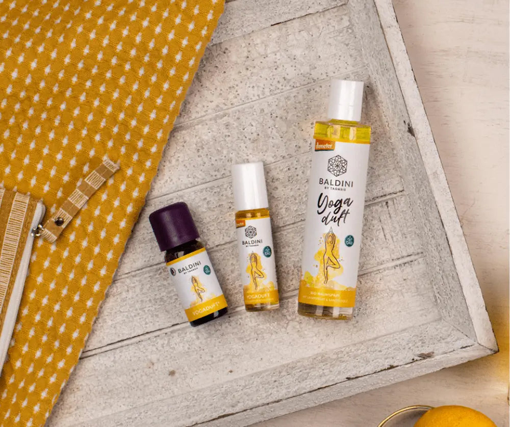 Drei Baldini Yoga Duft Produkte von Taoasis auf weißem Holztablett: Bio-Raumspray, Roll-on und Aromaöl-Flasche mit gelbem Yoga-Motiv